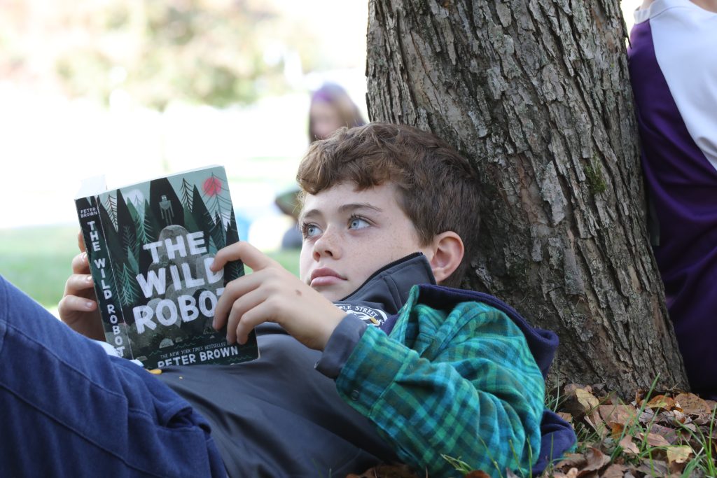 An older Lower School student rests against a tree trunk, reading a book called The Wild Robot.